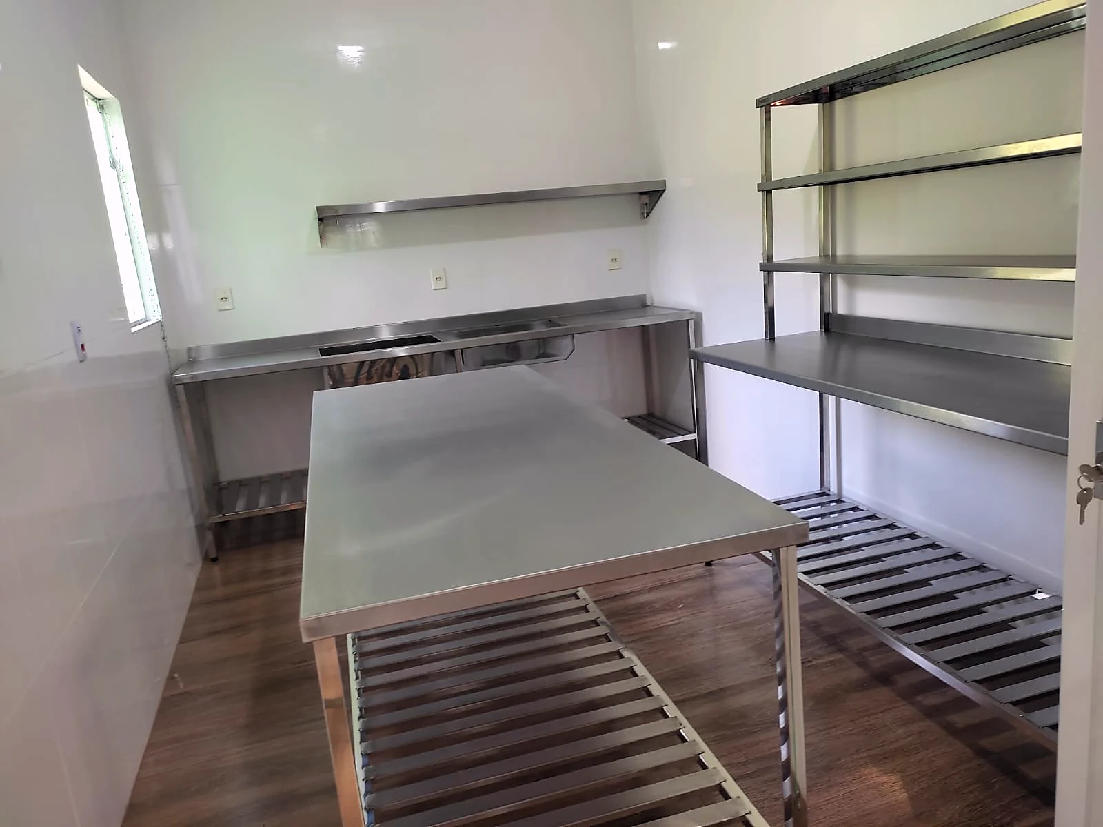 Cozinha industrial montada com bancada encostada na parede, mesa central de apoio e estante em inox