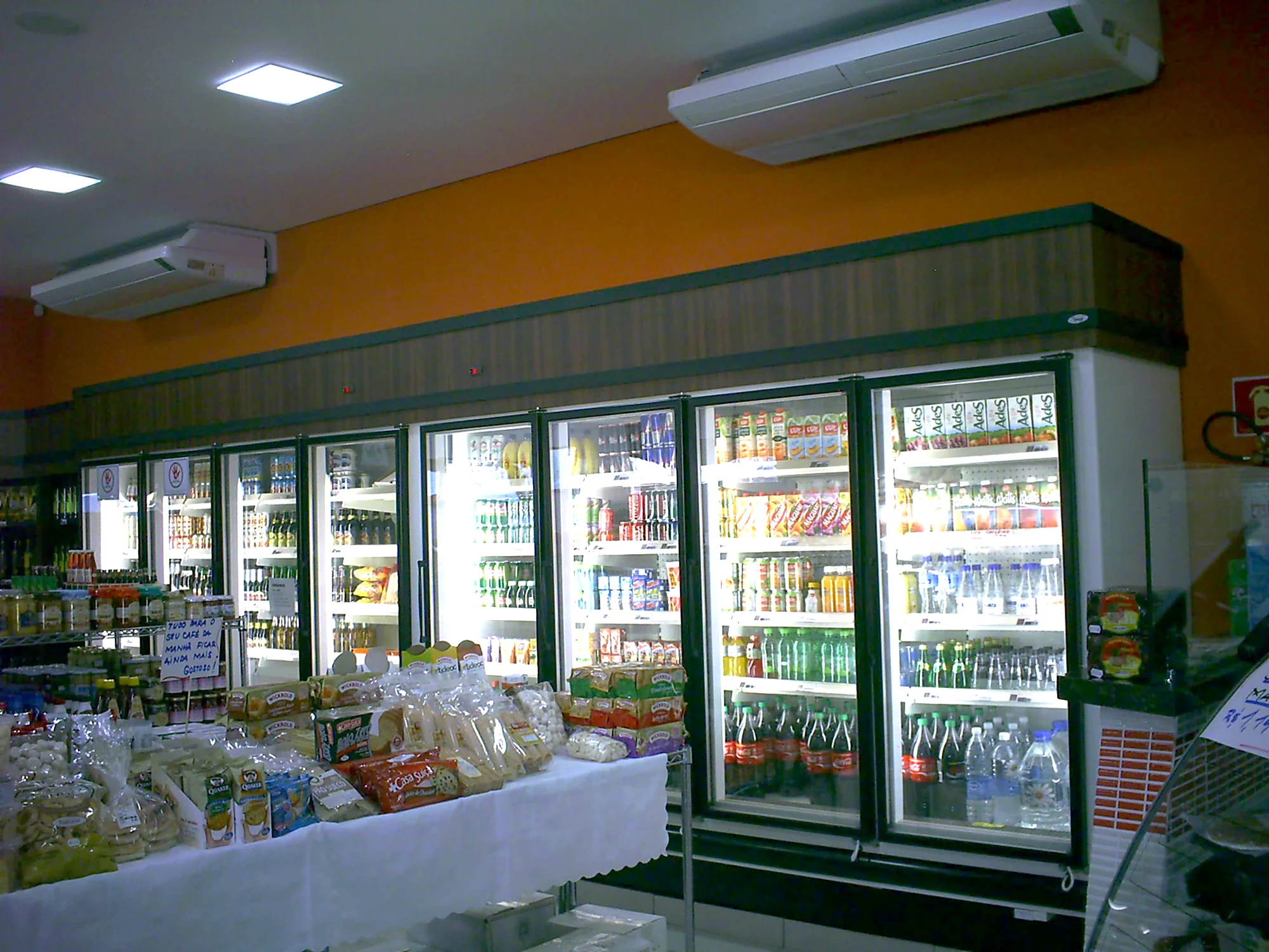 Linha contínua de refrigeradores verticais com portas de vidro, iluminação interna e prateleiras abastecidas de bebidas em um mercado