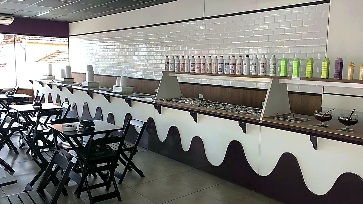 Balcão de atendimento de sorveteria com frente branca ondulada, tampo corrido e banquetas ao longo do salão